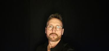 Man with glasses and beard against black background.