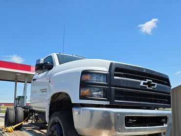 White Chevrolet Silverado 6500 HD truck secured on a flatbed trailer at a gas station.