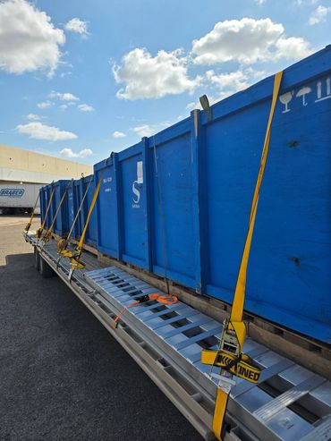 Blue wooden crates secured with yellow straps on a flatbed trailer under a partly cloudy sky.
