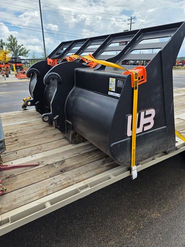 Three large black loader buckets secured on a wooden trailer bed.