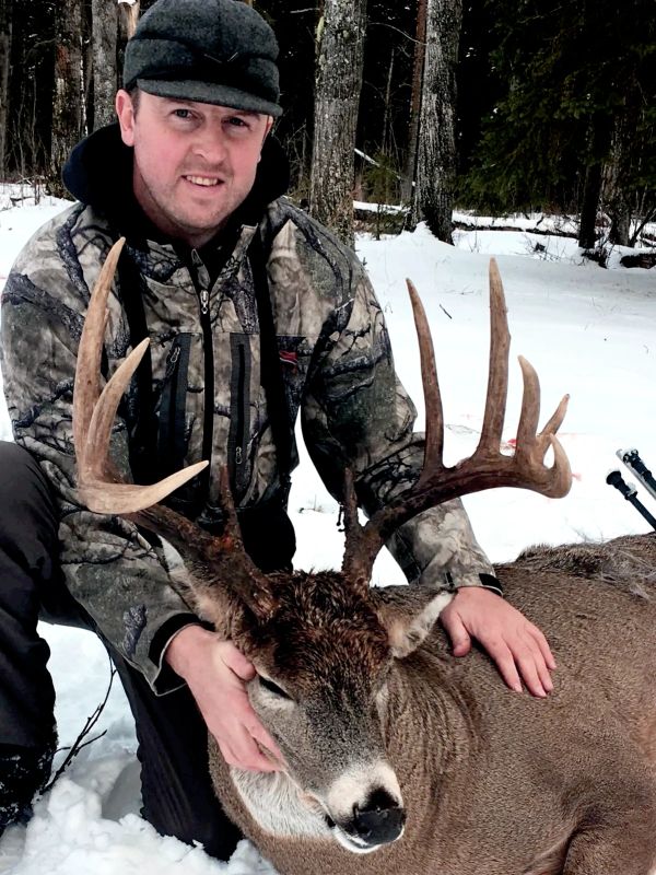 Hunter posing with a large buck deer in snowy forest.