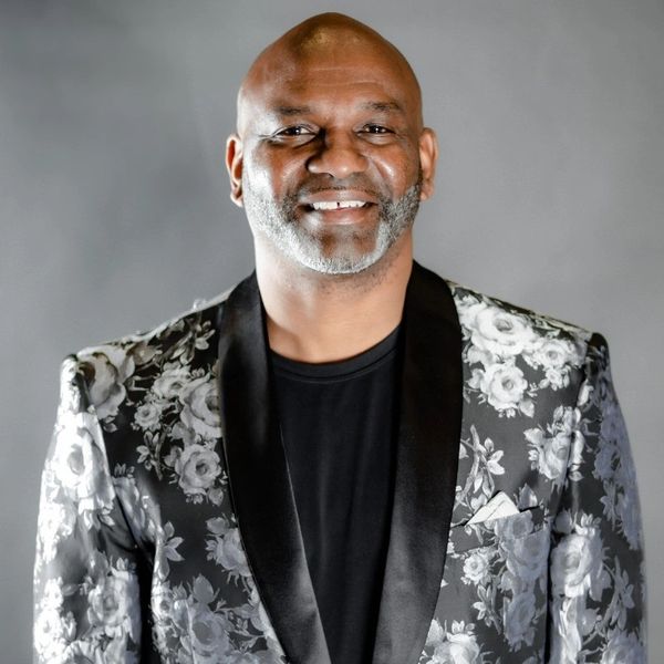 Smiling man in a black floral-patterned blazer and black shirt.