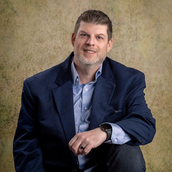 Man in a navy blazer and light blue shirt posing against a textured backdrop.