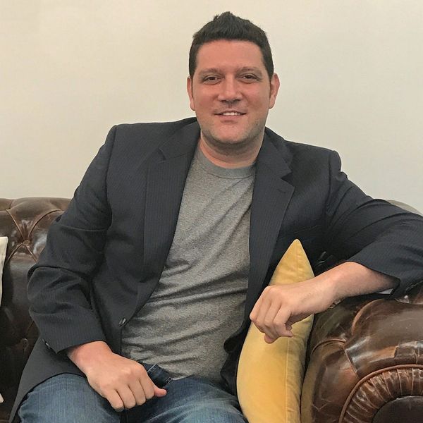 Man in a blazer and jeans sitting on a brown leather couch with a yellow pillow.
