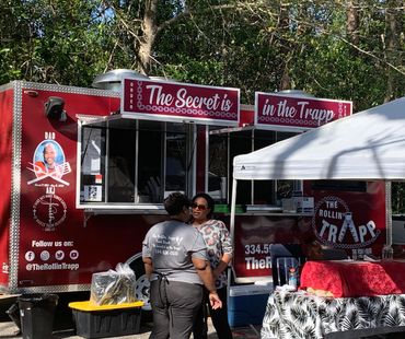 Two women chatting near a red food truck named The Rollin' Trapp in a wooded outdoor setting.