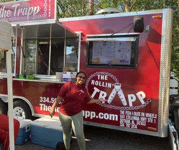Smiling woman posing with The Rollin' Trapp food truck in a sunny outdoor setting.