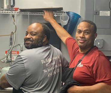 Two men smiling in a kitchen, one wearing an apron and the other a gray shirt.
