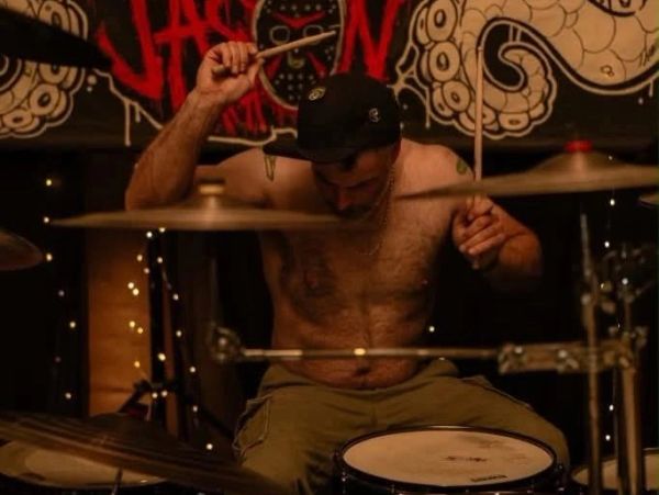 Shirtless man passionately playing drums in a dimly lit setting.