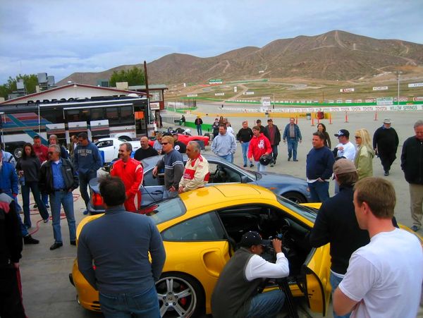 A large group of people gathered around several cars in the pits of the track