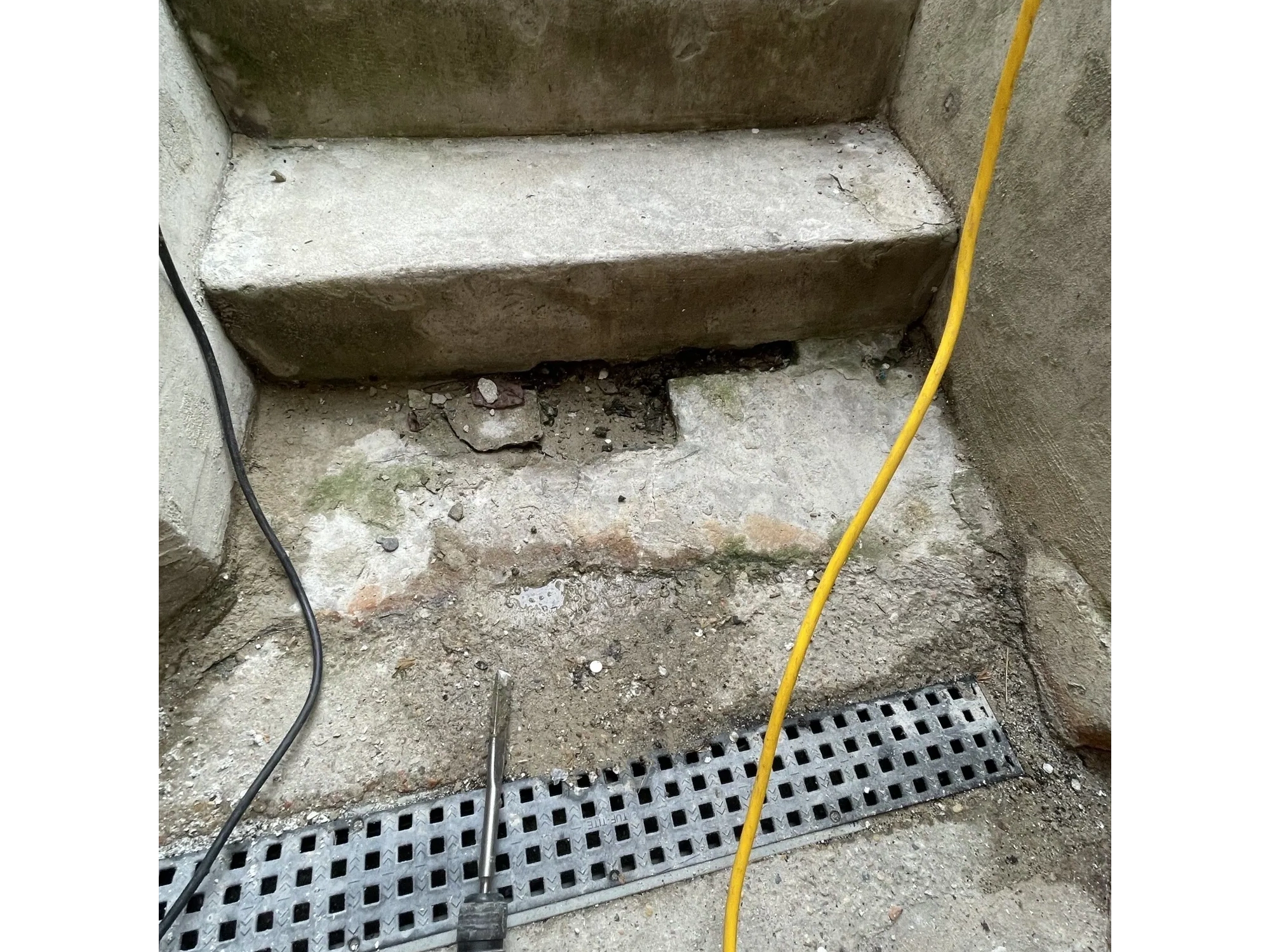 Damaged concrete stair landing with drainage grate, showing broken concrete and surface deterioratio