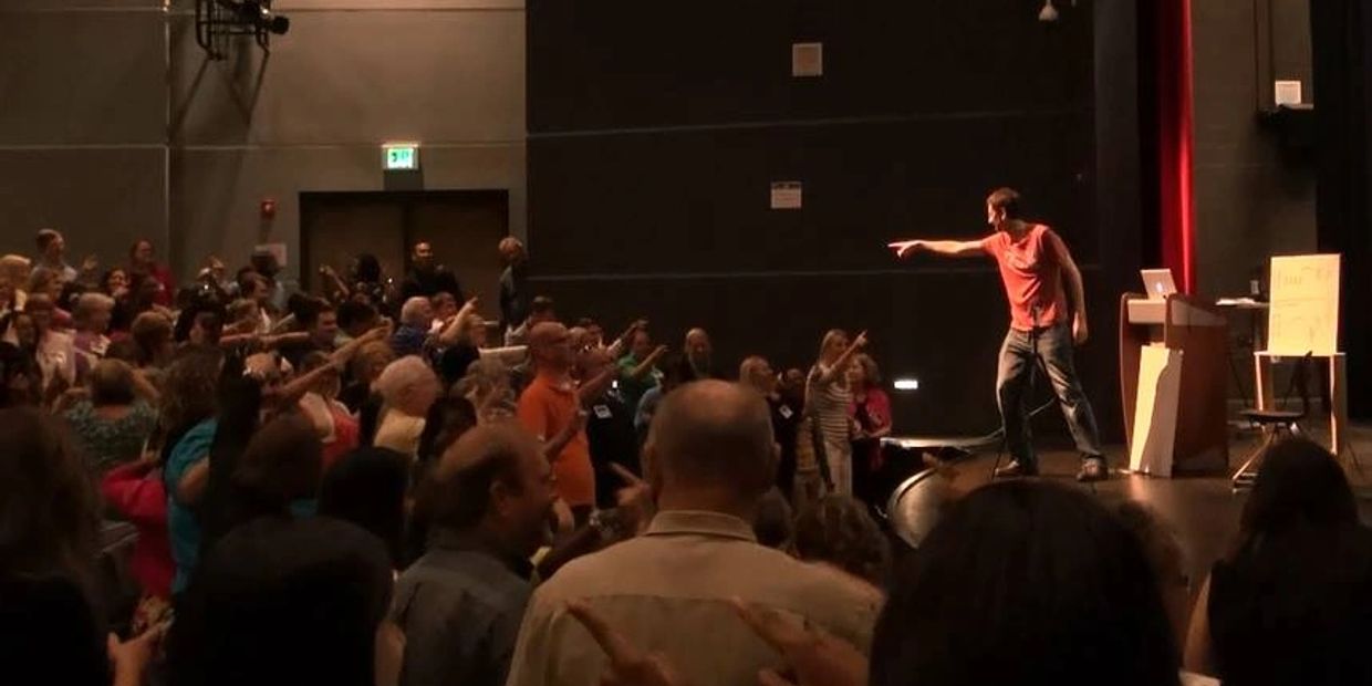 A speaker points at an audience in a dark auditorium.