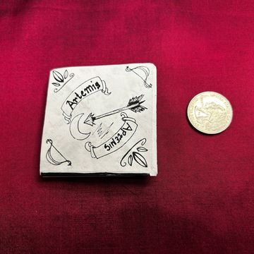Hand-drawn Artemis and archery design on a small book cover beside a coin.