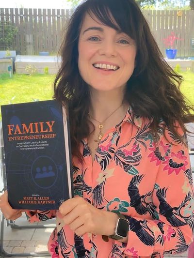 A picture of Prof McAdam in a pink patterned blouse smiling and holding the book Family Entrepreneur