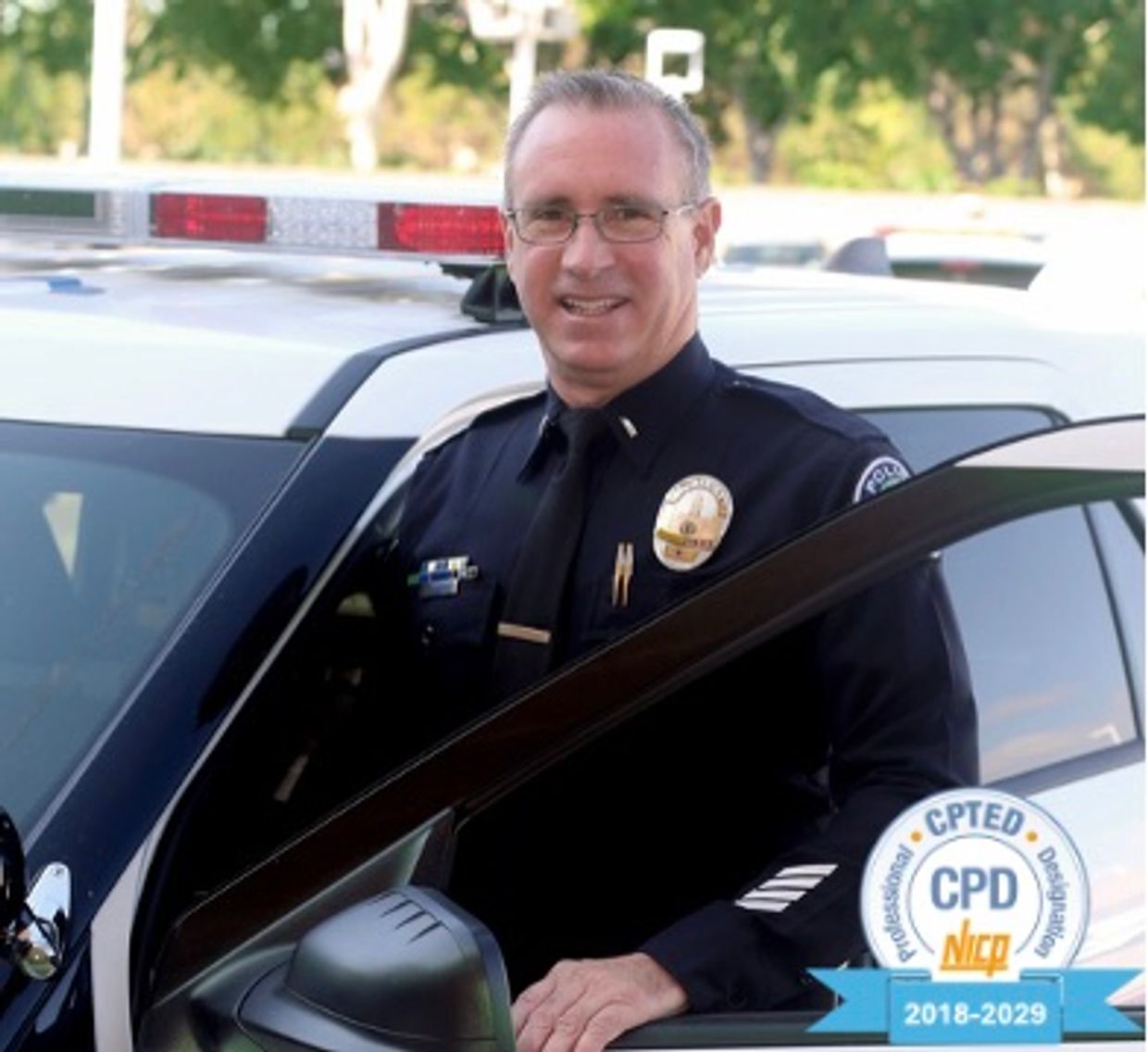 A smiling police officer standing by a patrol car with a CPTED certification badge.