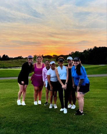 Seven women posing on a golf course at sunset, smiling and dressed in sporty golf attire.