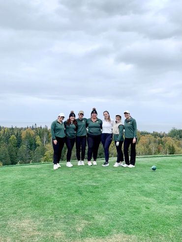 Group of seven women posing on a golf course with a forest backdrop under a cloudy sky.