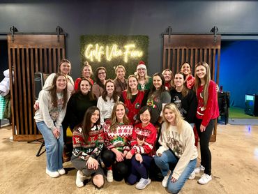 A group of women in festive sweaters posing together indoors under a "Golf Vibe Fun" neon sign.