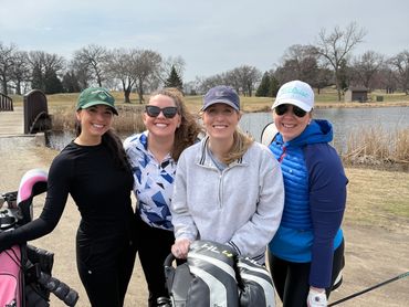 Four women posing with golf gear on a cloudy day at a golf course.