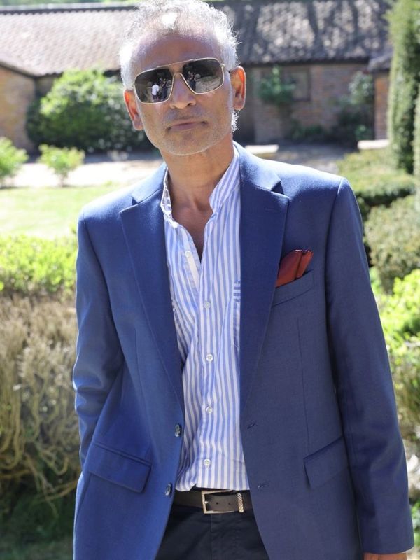 Man wearing sunglasses and a blue blazer outdoors in a garden.