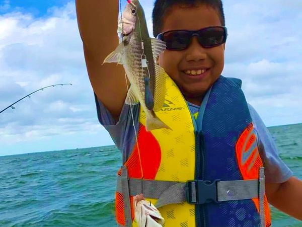 A boy wearing a life jacket proudly holds two fish on a boat.