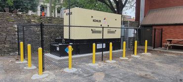 A large Kohler diesel generator enclosed by a chain-link fence with yellow bollards.