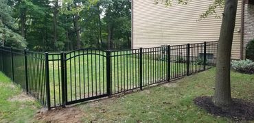 Black metal fence enclosing a green yard beside a beige house with an arched gate.