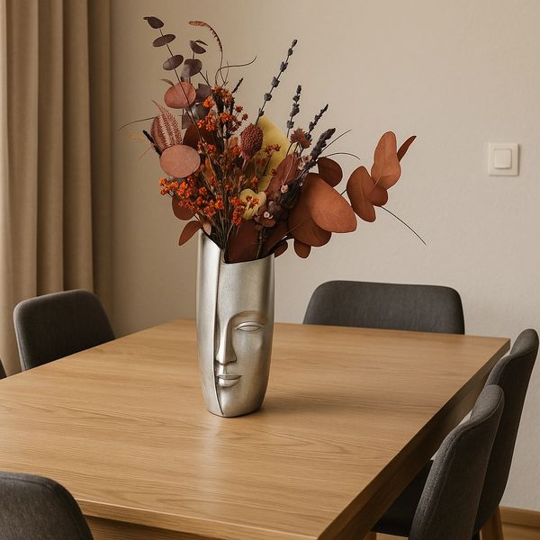 Silver vase with human face on its front, holding a bouquet of preserved foliage in autumnal tones