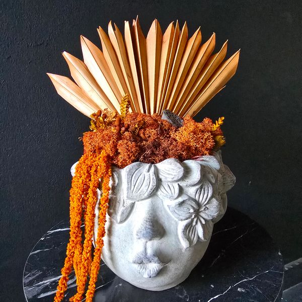 Head-shaped planter is filled with textured orange preserved moss, dried orange palm leaves.
