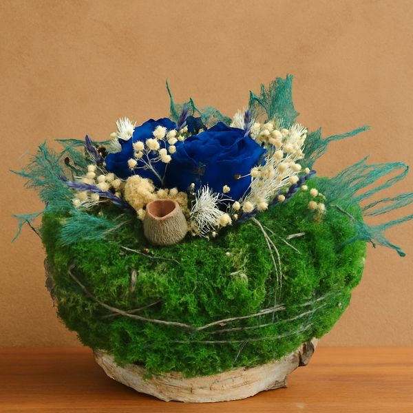 A circular preserved floral arrangement blue roses and green moss, on a natural wood slice base