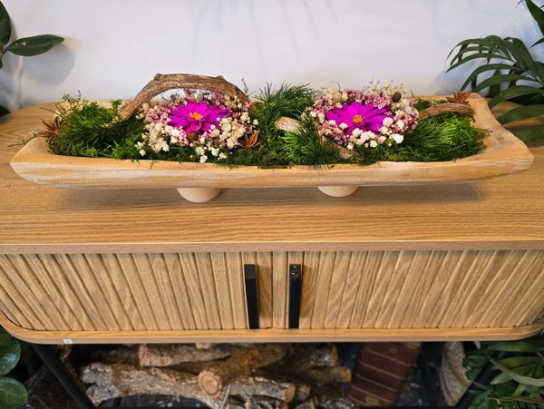 A long, rectangular wooden planter filled with moss, flowers, and branches sits on a wooden cabinet.