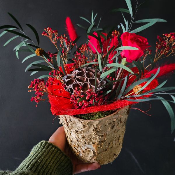 A rustic birch bark pot holds an arrangement of preserved red roses, and dark green leaves.