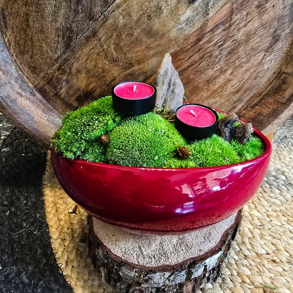 A red bowl contains green preserved moss, two small black candle holders with candles