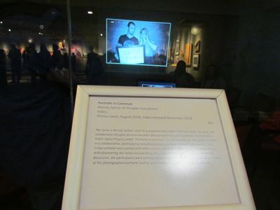 Gallery note in foreground explains project, and screen behind it, in darkened room, projects video.