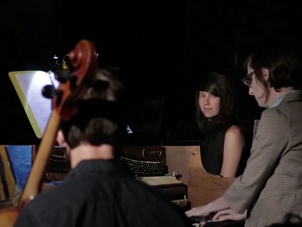 Woman looks at second woman who is at a piano; cellist in foreground. They are performing live.