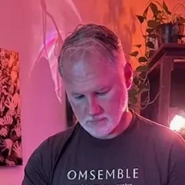Man with gray hair and beard, wearing a black shirt, illuminated by pink and orange light.