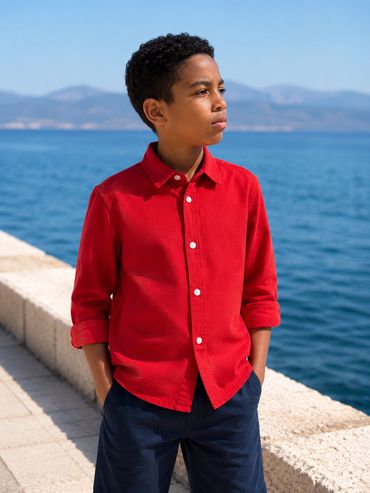 Young boy in red shirt standing by the sea, looking thoughtfully into the distance.
