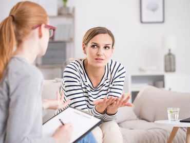 A woman on a couch gestures while speaking to another person holding a clipboard.