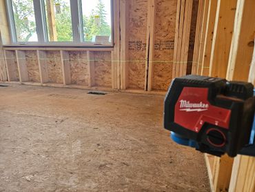 A Milwaukee laser level projecting a green line in a wooden-framed room under construction.