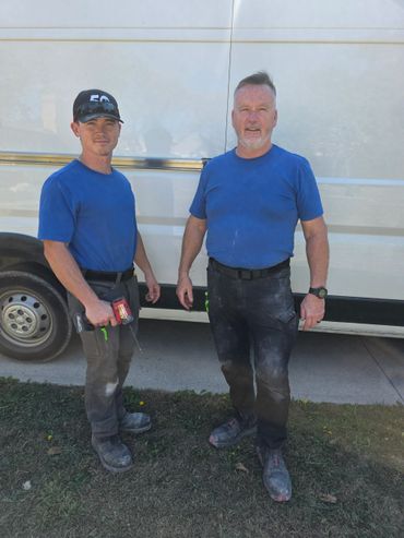 Two construction workers in blue shirts standing by a white van.
