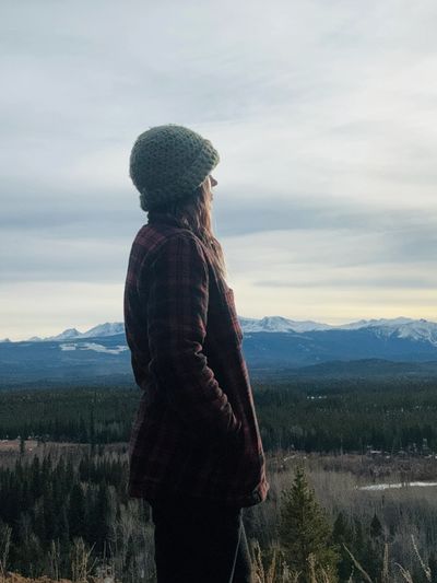 Person in a knit hat gazes over a vast forest and mountain landscape.