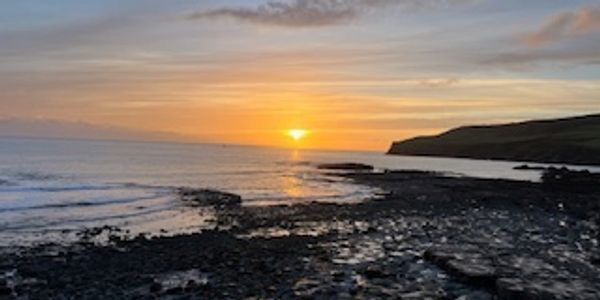 Sunset over a rocky shoreline with calm sea and distant hills.