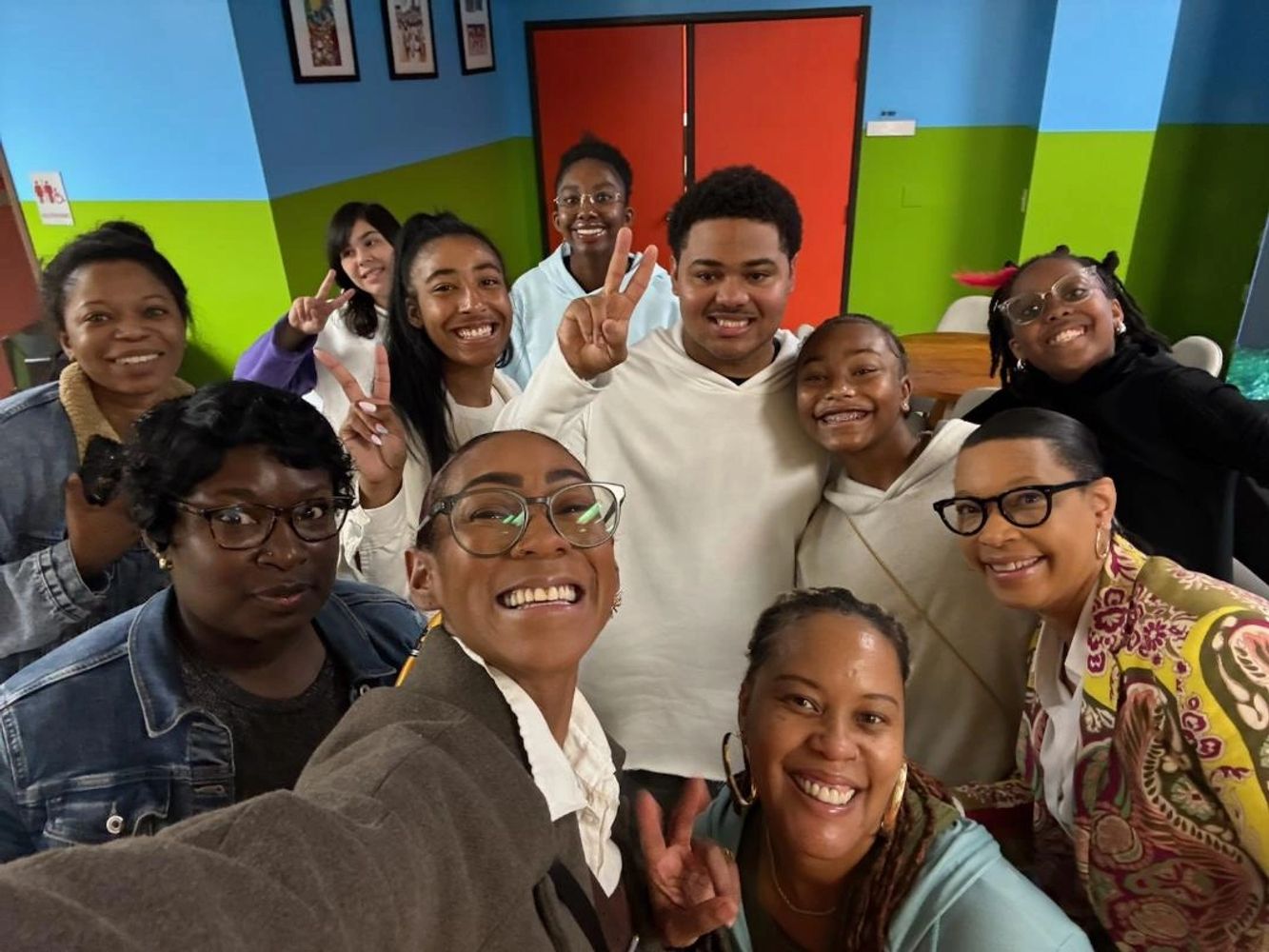 A joyful group selfie with diverse people showing peace signs and big smiles indoors.