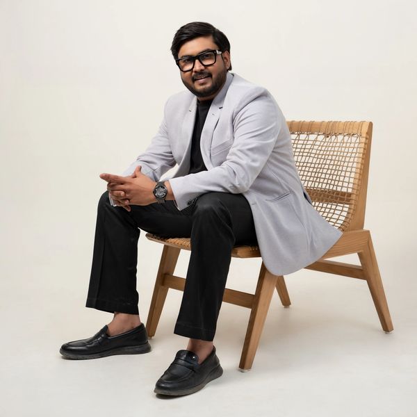 Man in a light gray blazer and black pants sitting on a modern wooden chair, smiling.