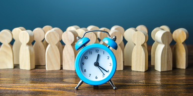 Blue alarm clock in front of wooden human figures on a table.