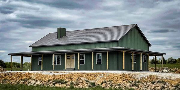 Newly constructed green pole barn with wraparound porch under cloudy sky.
