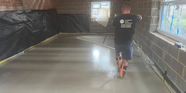 Worker smoothing concrete floor in unfinished room with exposed beams.