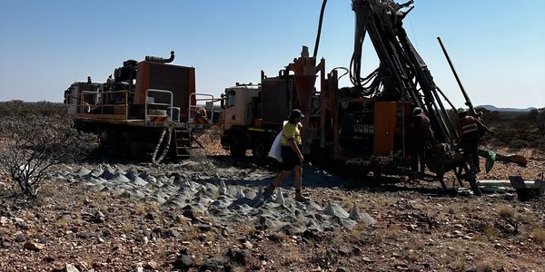 Workers operate heavy drilling machinery in a rocky, arid landscape under a clear blue sky.
