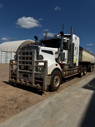 White Kenworth semi-truck with a bull bar parked on dirt under a blue sky.