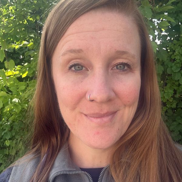 Close-up of a smiling woman with light brown hair and a nose ring outdoors.