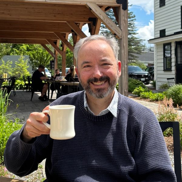 Man smiling and holding a white mug outdoors at a café.